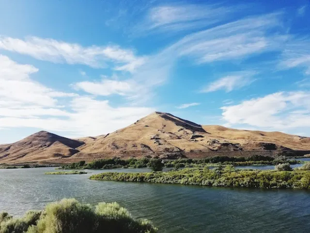 Scenic view of Deer Flat National Wildlife Refuge with Snake River, green vegetation, and rolling hills under a blue sky in Nampa, Idaho
